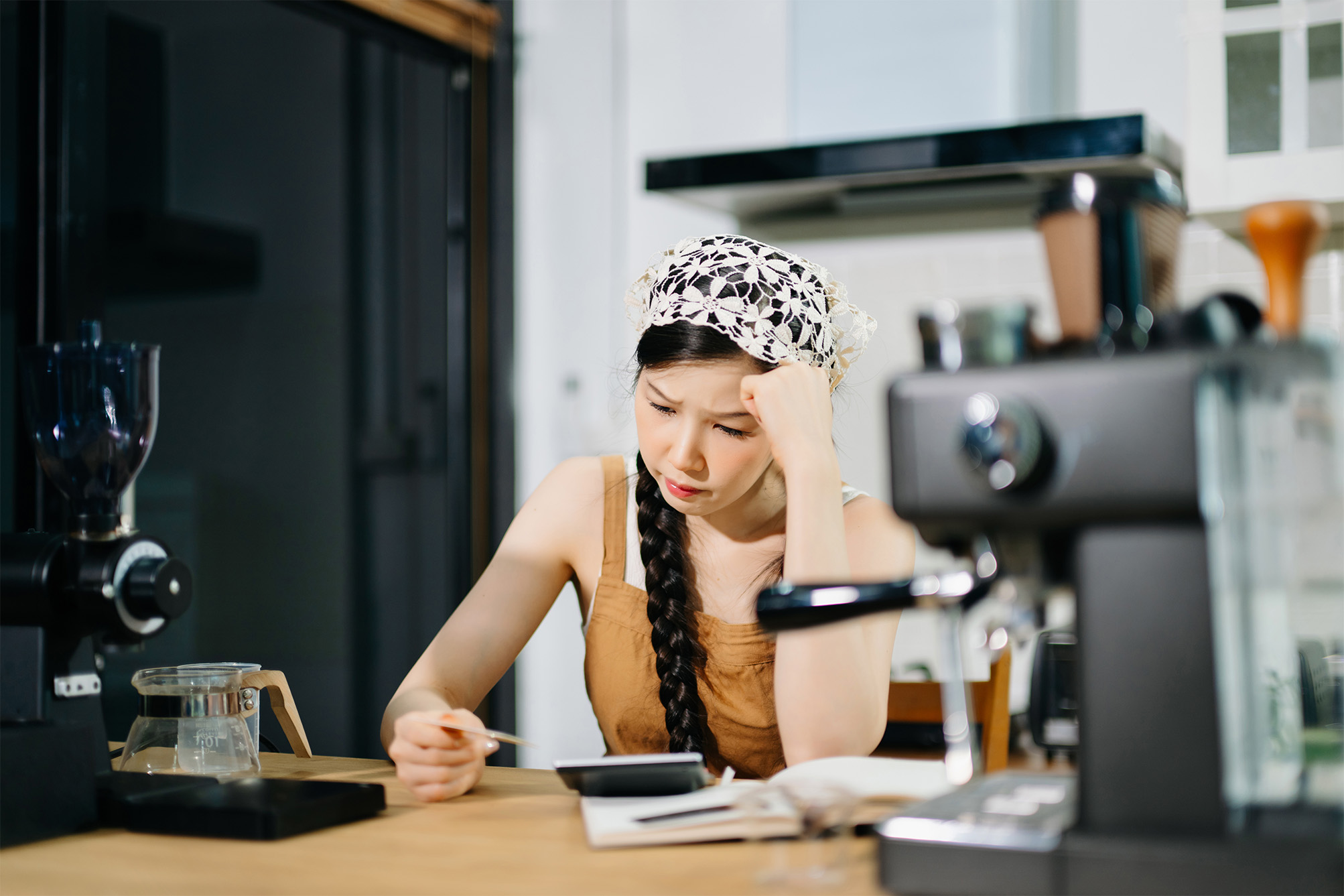 A stressed New York café owner reviewing costs with a calculator and notebook at her counter.